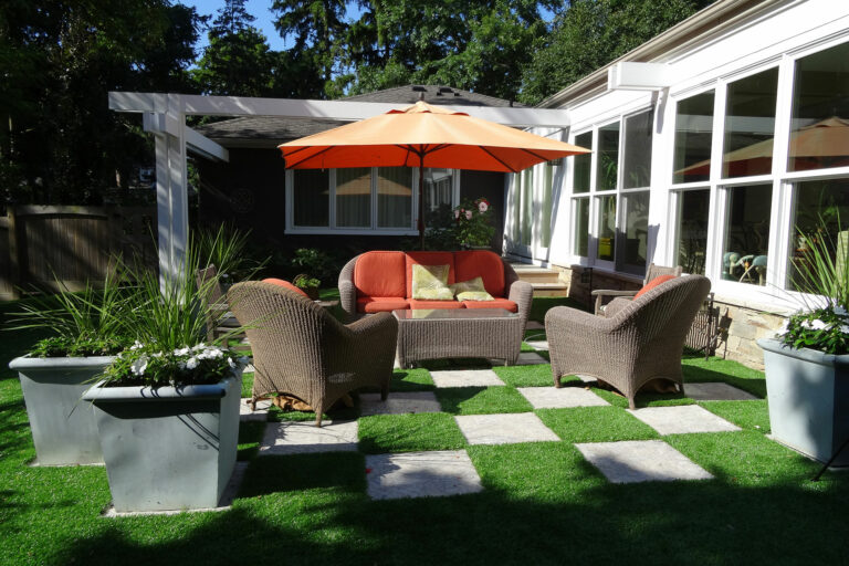 Outdoor seating set on checker-pattern synthetic grass and stone pavers, shaded by orange patio umbrella.