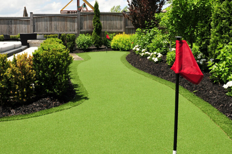 Curved putting green bordered with synthetic grass and red flag, surrounded by mulch and flowering bushes.