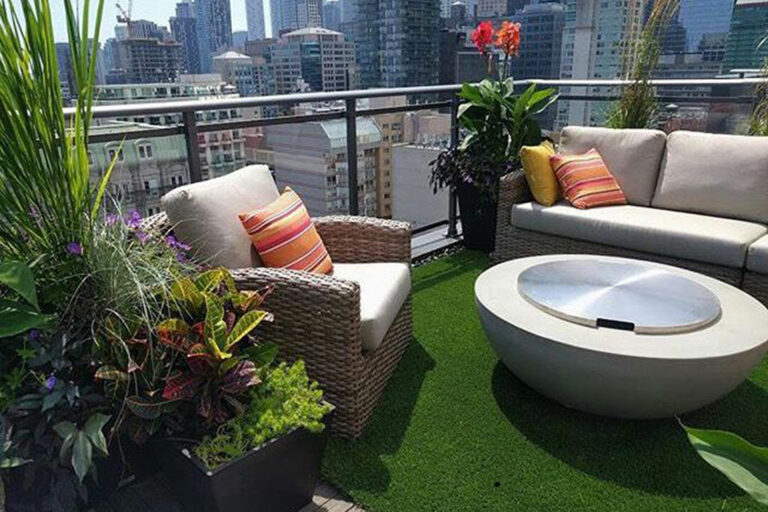 Cozy balcony with wicker chairs, cushions, and firepit on synthetic grass, surrounded by tall buildings.
