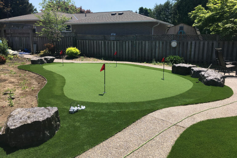 Private backyard putting green with short synthetic grass, red flags, and rocks lining the curved border.