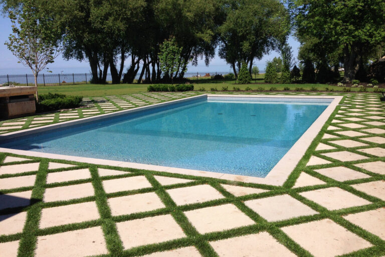 Outdoor pool surrounded by square tiles with synthetic grass lines, set in large lawn near wooded lakefront.